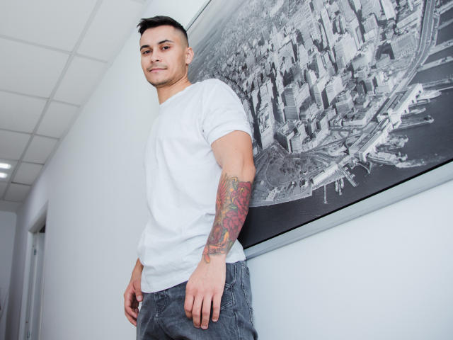 MattMalone, met een wit T-shirt, blauwe jeans en een getatoeëerde arm, staat in een gang en kijkt met een lichte glimlach naar de camera. Op de muur hangt een zwart-wit foto van een stad..