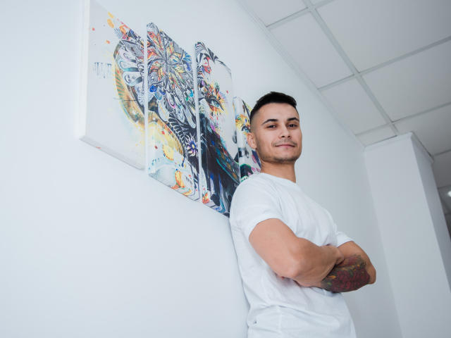 MattMalone, standing with arms crossed, wearing a white t-shirt, smiling, short dark hair, tattoo on right forearm, white wall with colorful abstract painting behind him.
