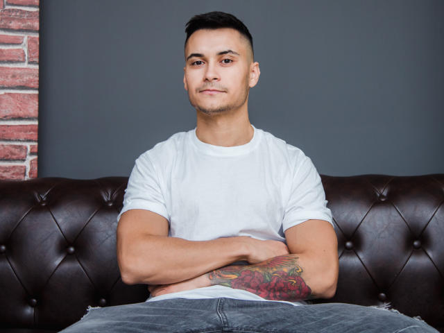 MattMalone, vestindo uma camiseta branca e calças jeans azuis, está sentado em um sofá de couro marrom, com os braços cruzados, uma tatuagem no antebraço direito, cabelo preto curto e um leve sorriso, com um fundo de parede cinza..