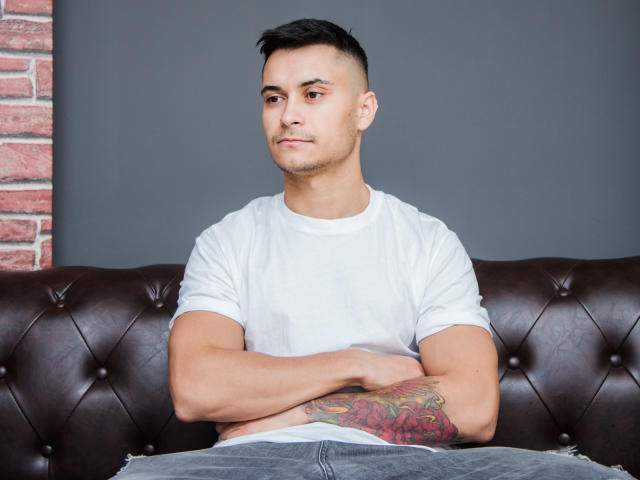 MattMalone mit einem Tattoo auf seinem rechten Unterarm, der ein weißes T-Shirt und blaue Jeans trägt, sitzt auf einem braunen, mit Leder bezogenen Sofa, die Arme verschränkt, mit kurzen, schwarzen Haaren..