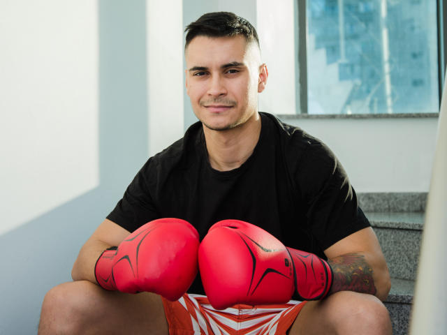 MattMalone, musculoso, de piel bronceada, cabello negro corto, camiseta negra, guantes de boxeo rojos, tatuaje en el brazo derecho, sentado en las escaleras, sonriendo, usando pantalones cortos a rayas naranja y blanco..