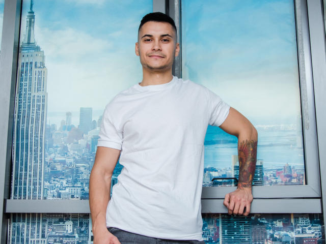 MattMalone se tient devant une fenêtre, vêtu d'un t-shirt blanc et d'un pantalon gris, avec un bras tatoué, des cheveux courts et foncés, et un léger sourire. Sa main repose sur la fenêtre..