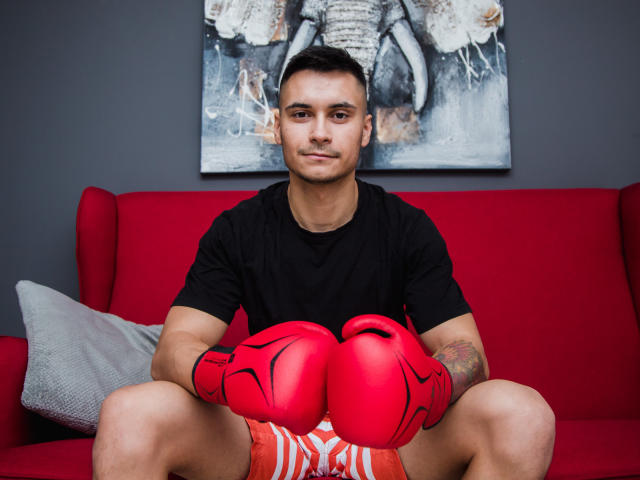 MattMalone, der rote Boxhandschuhe, ein schwarzes T-Shirt und rote Shorts trägt, sitzt auf einem roten Sofa mit einem grauen Kissen, die Arme auf den Knien ruhend, und schaut in die Kamera..