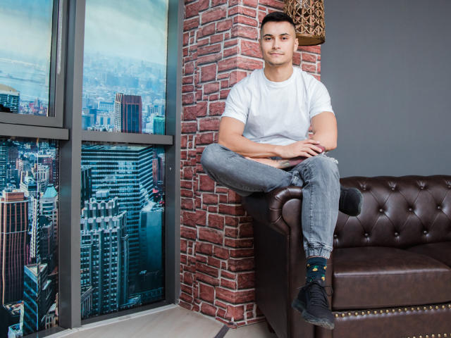 MattMalone sitting on a brown leather couch, wearing a white t-shirt, grey jeans, and black socks with yellow stars, hands on knees, brick wall and cityscape window behind.