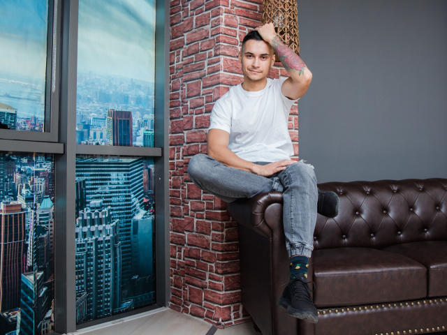 MattMalone, sentado en un sofá de cuero marrón, con camiseta blanca, jeans azules, calcetines negros, una mano en el pelo, una pared de ladrillo, vista de la ciudad a través de la ventana, un tatuaje en el brazo derecho, sonriendo..