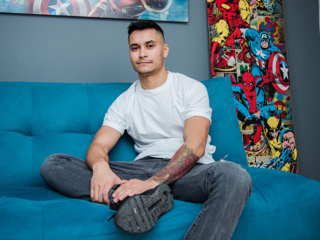 MattMalone, sitting on a blue couch, wearing a white tee, grey jeans, and black shoes, with a colorful tattoo on his right arm, looking at the camera.