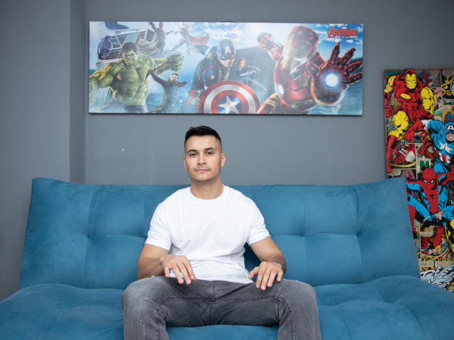 MattMalone, gespierd, kort zwart haar, wit T-shirt, grijze jeans, zittend op een blauwe bank, schilderij van de Avengers erboven, kleurrijk superhelden schilderij aan de rechterkant, neutrale uitdrukking, kleine ring aan de rechterhand..