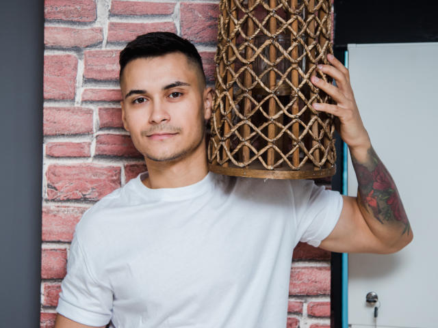 MattMalone tenant une lampe en osier avec un motif géométrique, portant un t-shirt blanc, avec des cheveux courts et foncés, un tatouage sur le bras droit, un léger sourire, debout devant un mur de briques..