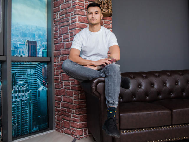 MattMalone with muscular arms, sitting on a brown leather couch, wearing a white tee, grey jeans, and black socks, looking serious, brick wall behind him, city view window.