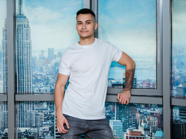 MattMalone, con una camiseta blanca, pantalones grises y tatuajes en su brazo derecho, apoyado contra una ventana con vistas a la ciudad, sonriendo ligeramente, con cabello negro corto y piel clara..