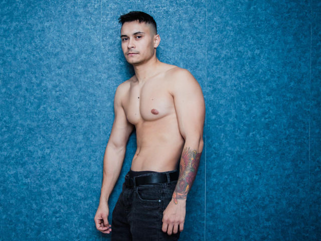 MattMalone, zonder shirt, met een getatoeëerde arm, zwarte jeans, staand tegen een blauwe, gestructureerde muur, kijkend iets opzij, kort zwart haar, gemiddeld postuur, lichte glimlach..