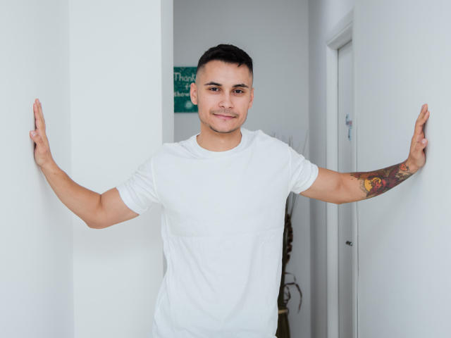 MattMalone con los brazos apoyados contra paredes blancas, vistiendo una camiseta blanca, cabello negro corto, tatuaje en el brazo derecho, sonriendo, de pie en un pasillo estrecho..