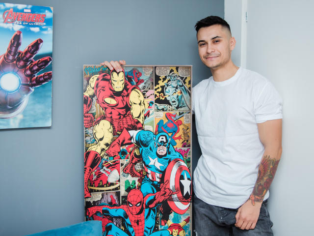 MattMalone, stehend, weißes T-Shirt, tätowierter Arm, kurze, dunkle Haare, lächelnd, lehnt sich an einen farbenfrohen Comic-Poster mit Marvel-Superhelden, darunter Spider-Man und Captain America..
