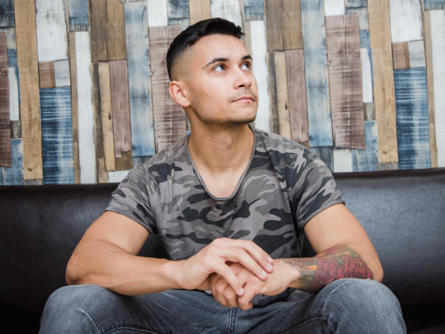 MattMalone, muscular, tan skin, short black hair, camo t-shirt, tattoo on right forearm, sitting on black leather couch, looking to the side, hands on knees, blue jeans.