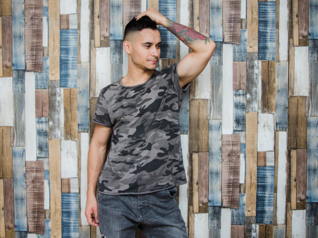 MattMalone è in piedi, con una mano sulla testa, indossa una maglietta grigia a fantasia camouflage e jeans blu, mostrando un braccio tatuato, e guarda di lato con un leggero sorriso..