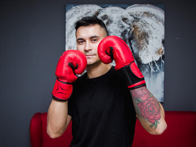 MattMalone, con i guantoni da boxe rossi, la maglietta nera e il braccio tatuato, in piedi di fronte a un dipinto, sorride leggermente, con le mani alzate in posizione da pugilato..