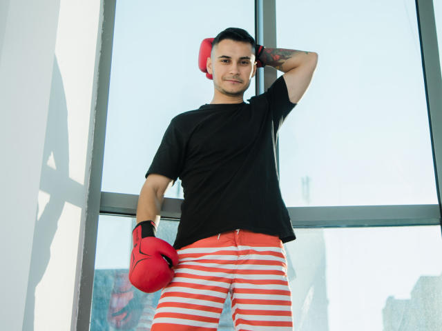 MattMalone, com cabelo preto curto, vestindo uma camiseta preta, luvas de boxe vermelhas e calças com listras vermelhas, está em frente a uma janela, com a mão atrás da cabeça, e parece confiante..