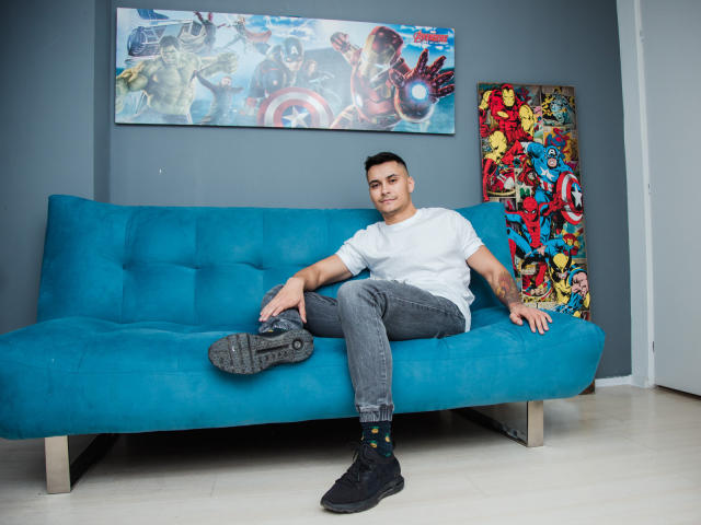 MattMalone sitzt auf einem blauen Sofa, trägt ein weißes T-Shirt, graue Jeans und schwarze Turnschuhe. Auf seinem rechten Arm sind Tattoos zu sehen. Er lächelt in die Kamera..