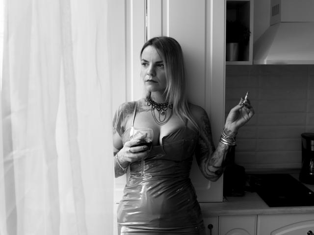 VanessaOdette staat in een keuken, gekleed in een glanzende latexjurk, met een glas rode wijn in haar hand, waarbij tatoeages op haar armen zichtbaar zijn, en een sigaret in haar rechterhand..