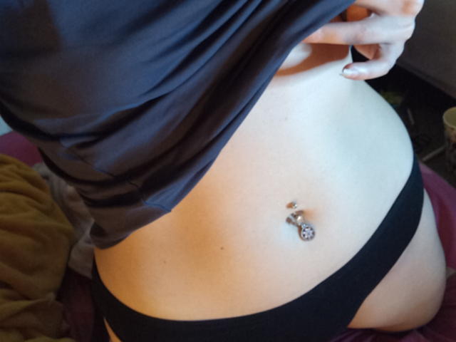 A barriga nua de VirginieFrancaise, com um piercing de prata no umbigo, calcinhas pretas e uma camisa cinza levantada, mostrando sua pele clara e macia, e levemente curvilínea e lisa..