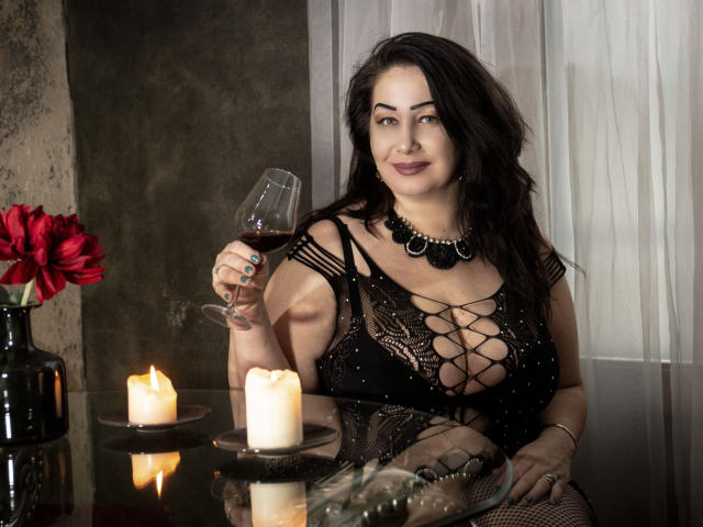 LexyRose with dark hair, wearing black lace lingerie, holding a glass of red wine, sitting by candles, red flower in vase, black necklace, smiling, large boobs, curvy.