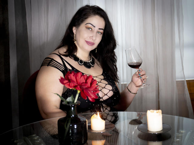 LexyRose with big tits, holding a glass of red wine, wearing a black dress with fishnet sleeves, sitting at a table with candles and a red flower.