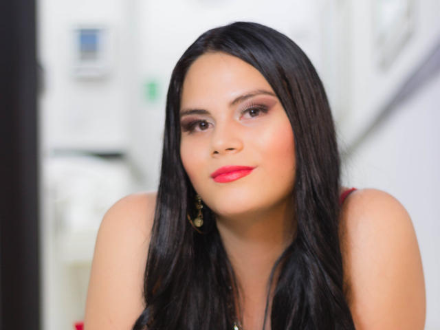 JassmineTS tem cabelos pretos longos, batom vermelho e está olhando para frente com um leve sorriso. Ela está usando brincos dourados e tem um tom de pele claro. Os seios estão parcialmente visíveis..