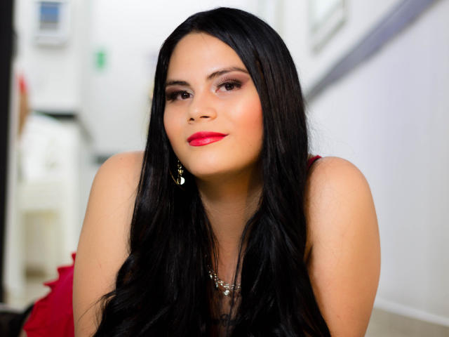 JassmineTS tem cabelos pretos longos, batom vermelho e um olhar confiante. Ela está usando um colar e um vestido vermelho, com maquiagem discreta e brincos dourados. Ela está olhando ligeiramente para o lado..