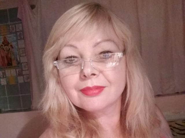 EvelynLoveShy, avec ses cheveux blonds, ses lunettes claires et son rouge à lèvres rouge, souriant légèrement, montrant son nez et ses lèvres, avec ses seins à peine visibles dans la partie inférieure du cadre..