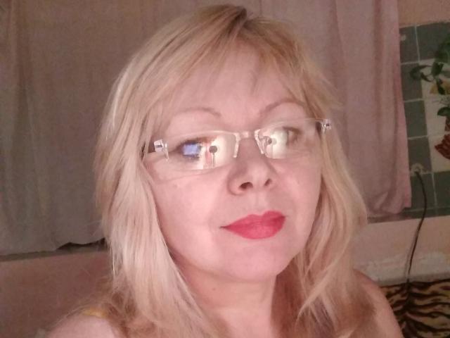 EvelynLoveShy tem cabelo loiro, óculos e batom vermelho, com um leve sorriso, olhando para o lado, mostrando seu nariz e olhos, com um reflexo em seus óculos..