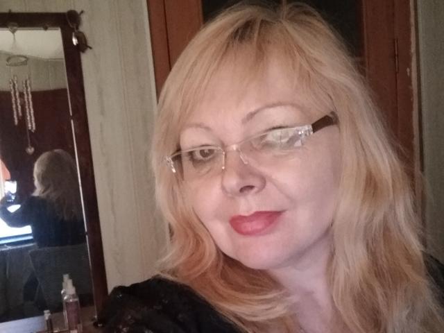 EvelynLoveShy a les cheveux blonds, porte des lunettes et du rouge à lèvres, et sourit avec le nez légèrement relevé. Son reflet dans le miroir montre son dos, un collier et une porte..