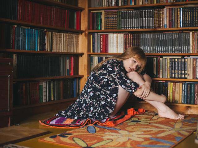 SweetAleksaX com cabelos vermelhos longos, sentada em um tapete colorido, vestindo um vestido floral preto, com os joelhos próximos ao peito, braços ao redor das pernas, em um quarto cheio de livros, descalça, com uma expressão triste..