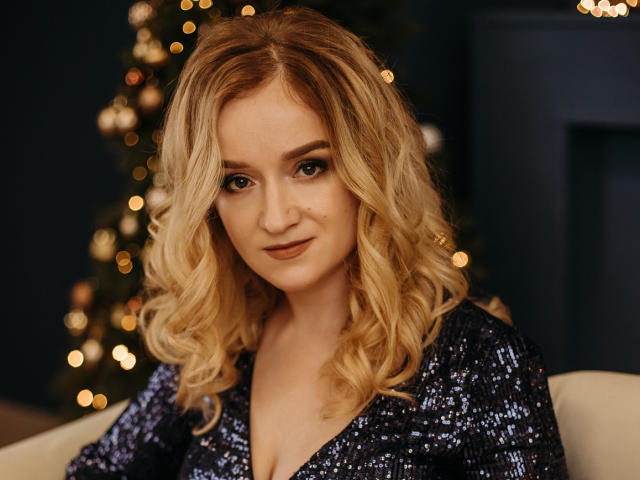OhMollyHoney with blonde curly hair, wearing a sparkly blue dress, has a cute face and a slight smile, sitting in front of a Christmas tree with warm lights.