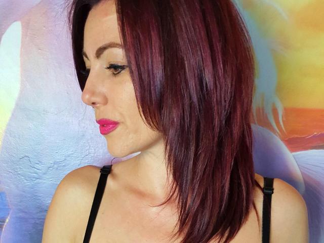 XKersX has red-purple hair, pink lipstick, black eyeliner, and is wearing a black strap top, showing a side profile with a slight smile. Boobs visible, no ass.