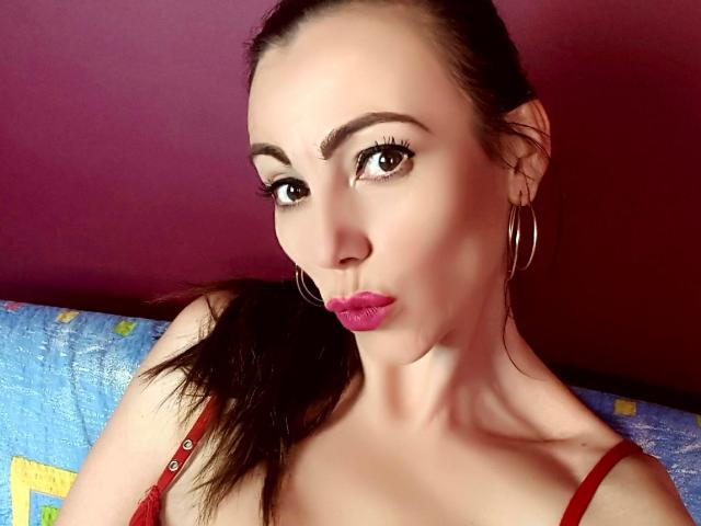 XKersX has pink lipstick, brown eyes, hoop earrings, and red lace top, with brown hair in a ponytail, making a pouty face. Her tits are partially visible.