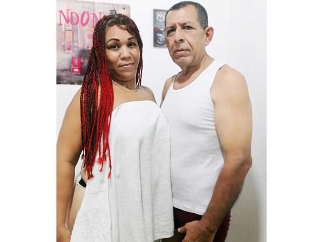 Pamela e Diego, com tranças vermelhas e uma toalha branca cobrindo os seios, estão em pé ao lado de um homem musculoso vestindo uma camiseta branca, ambos com uma expressão séria, em um pequeno quarto..