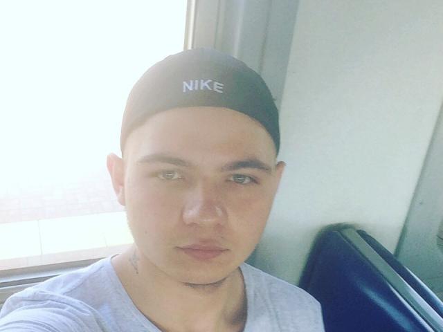 YarOGet, portant une casquette noire Nike et un t-shirt gris, a une expression sérieuse, une peau claire et des yeux verts, et se tient devant une fenêtre lumineuse..