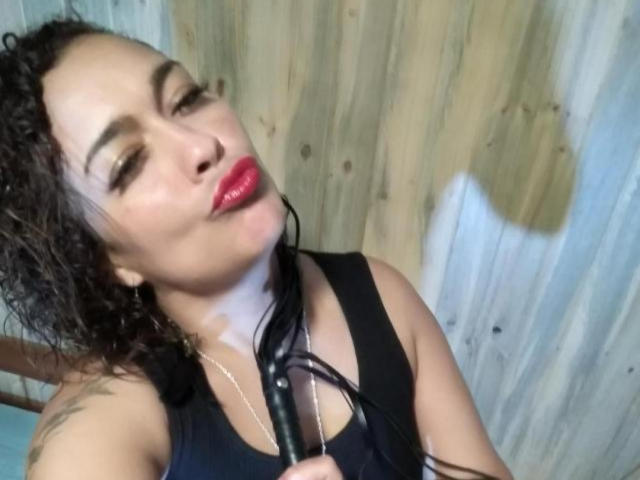 SolmaryHot with curly black hair, red lipstick, holding a black dildo against her chest, wearing a black tank top, wooden background, slight smile, visible tattoo on right shoulder.
