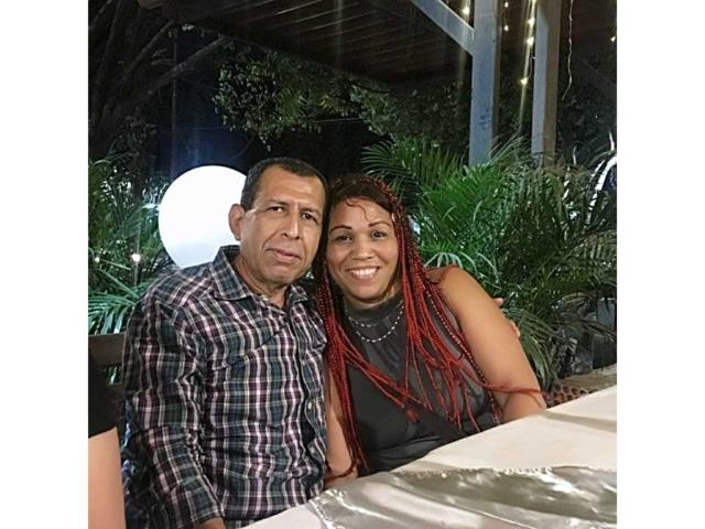 Pamela e Diego, com cabelo trançado ruivo, seios grandes, blusa preta sem mangas, e um homem de meia-idade sorrindo, vestindo uma camisa xadrez, em pé do lado de fora à noite..