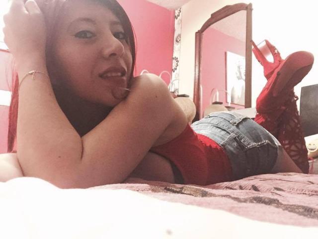 HotPamelaSex, laying on bed, red shirt, denim shorts, red boots, smiling, black hair, arm resting on forehead, looking at camera, pink bedroom, casual, sexy, relaxed.