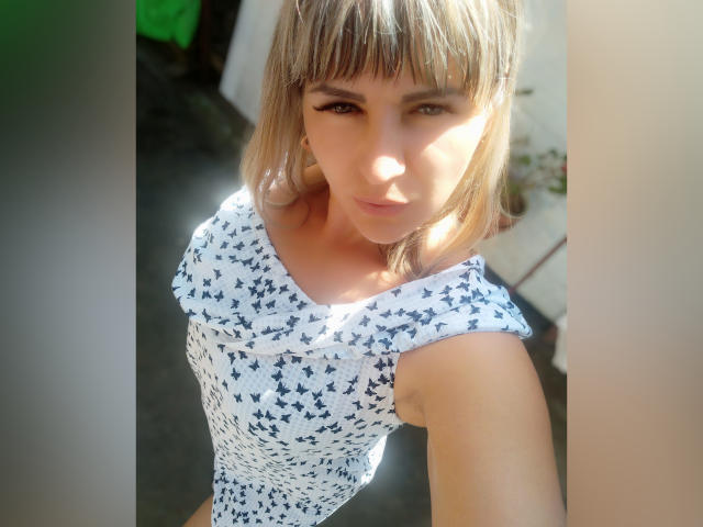 NancySkyblue, con cabello rubio corto, vestido azul con estampado de mariposas, piel clara, mirando hacia arriba, luz del sol en el rostro, pequeños senos, brazos delgados, sonrisa suave, linda, selfie realista, luz brillante al aire libre..