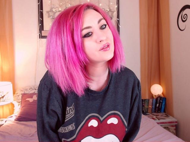 LuxNoir with pink hair, wearing a dark gray sweatshirt with a red mouth graphic, has a cute, slightly open-mouthed, pouty expression. Bedroom setting, soft lighting.