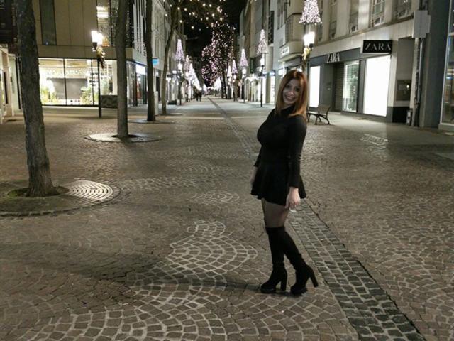 IssaDiamondd steht nachts auf einer Kopfsteinpflasterstraße, trägt ein schwarzes Langarmshirt, einen Minirock und kniehohe Stiefel und lächelt, wobei ihre Brüste leicht unter dem Oberteil sichtbar sind..