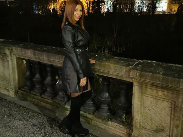 IssaDiamondd has brown hair, black leather dress with belt, black thigh-high boots, standing against a stone balcony at night, smiling, showing her ass, with trees in background.