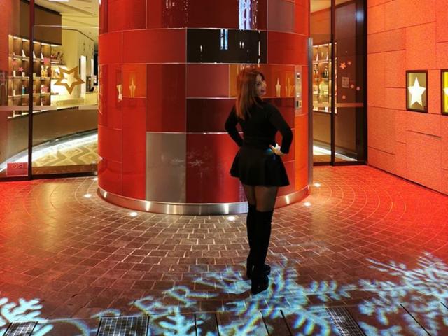 IssaDiamondd mit langen braunen Haaren, einem schwarzen Kleid und kniehohen Stiefeln, steht vor einer roten, zylindrischen Struktur und wirft weiße, schneeartige Schatten auf den gefliesten Boden..