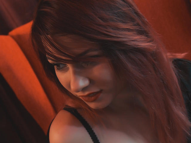 AliciaKristal mit rotbraunen Haaren, die nach unten schaut, in einem schwarzen Trägerkleid, mit einem leichten Lächeln, dunklem Lippenstift, weichem Licht, orangefarbenem Hintergrund, dezenter Make-up, sanftem Ausdruck, weichem Fokus, intime Aufnahme..