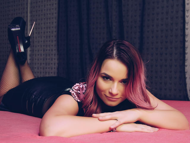 AliciaKristal, con i capelli rosa, sdraiata su un letto rosa, indossa pantaloni di pelle nera e collant a rete, tacchi alti neri, e guarda la fotocamera con un sorriso seducente..
