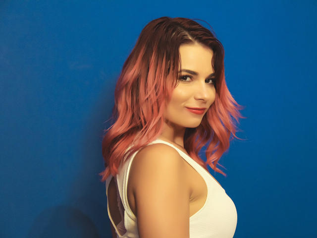 AliciaKristal mit rosa, gewellten Haaren, einem weißen Tanktop und einem verschmitzten Lächeln, steht vor einem blauen Hintergrund und zeigt ihr niedliches Hinterteil und ihre kleinen Brüste. Sie schaut über ihre Schulter..