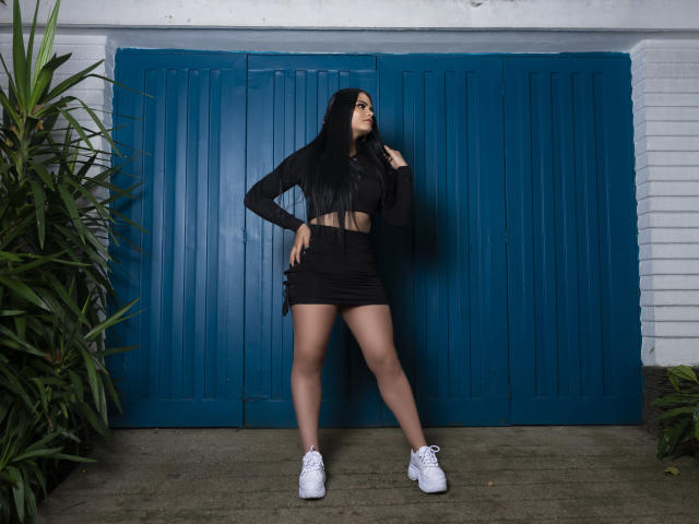 JassmineTS, avec ses longs cheveux noirs, son crop top noir et sa mini-jupe, se tient debout, une main sur la tête, portant des baskets blanches, devant des portes bleues, montrant ses fesses et son ventre..