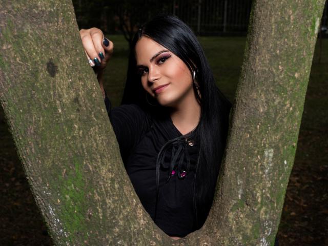 JassmineTS with black hair, wearing a black top, sticking her arm through a tree's V-shaped trunk, smiling slightly, with green fingernails and hoop earrings, in a dark setting.